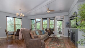 A spacious living room with a large sofa, a dining table with chairs, a fireplace, and large windows offering a view of the outdoors. - Best location in Salida! 2 River front decks, looks over Arkansas River waterpark! (Salida)