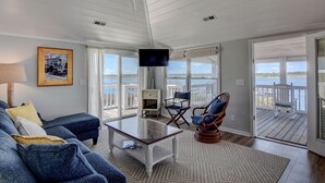Un salón con una gran mesa de vidrio, un sofá azul y un televisor. Tiene vista al agua y una terraza exterior.
