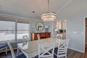 Uma sala de jantar com mesa de vidro e cadeiras brancas, um lustre e vista para o exterior através de grandes janelas.