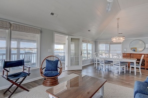 A spacious living room with a large window overlooking a body of water, a dining area with white chairs, and a wooden ceiling. - Harbor View–Enjoy the best sunsets on the island from this upper level duplex (Wrightsville Beach)