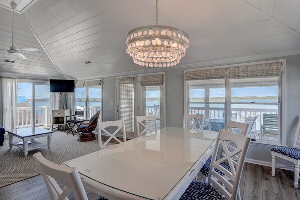 A spacious dining area with a large table and chairs, a chandelier, and large windows offering a view of the ocean. - Harbor View–Enjoy the best sunsets on the island from this upper level duplex (Wrightsville Beach)