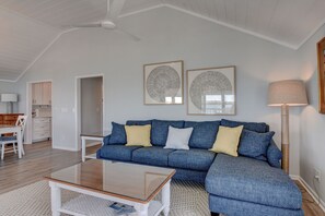 A living room with a blue sectional sofa, a glass coffee table, and two framed abstract artworks on the wall. - Harbor View–Enjoy the best sunsets on the island from this upper level duplex (Wrightsville Beach)