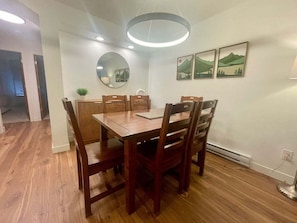 A dining area with a wooden table and chairs, a round mirror, recessed ceiling lights, and framed landscape paintings on the wall.