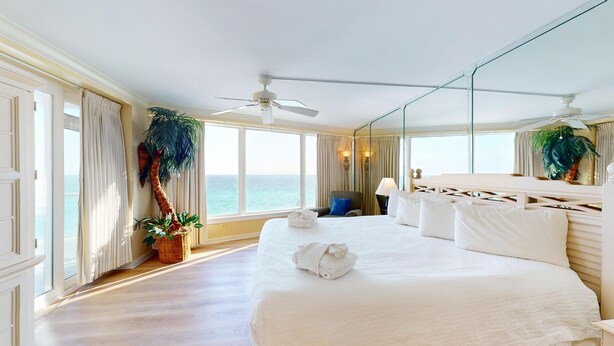 Habitación de hotel con una cama grande, ventilador de techo, vista al mar, una silla y una planta en maceta.