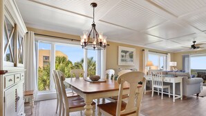 A spacious dining area with a wooden table and chairs, a chandelier, and large windows offering a view of palm trees. - Shorewood 312 -  3 bedroom Beautiful Ocean Front (Hilton Head Island)