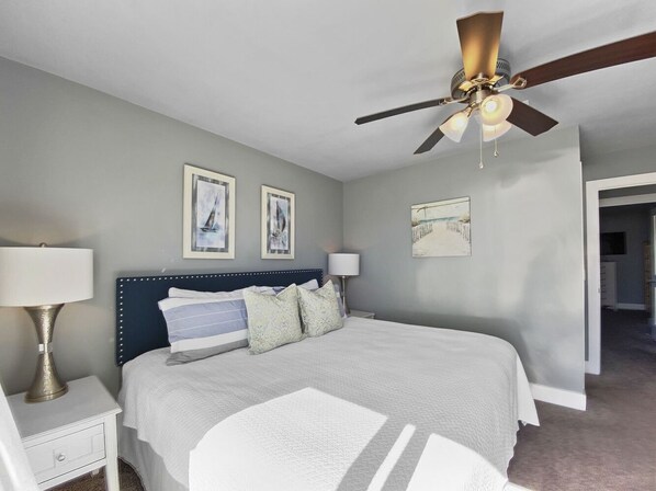 Une chambre à coucher avec un lit, une table de chevet, une lampe, un ventilateur de plafond et des tableaux encadrés accrochés au mur.