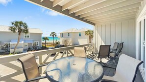 A covered outdoor seating area with a round table and chairs, overlooking a beach and neighboring houses.