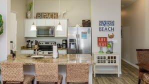A kitchen with granite countertops, white cabinetry, a stainless steel refrigerator, and a built-in microwave. There are wicker chairs around the kitchen island, and decorative signs on the walls. - Family & Dog-Friendly Townhome | 2 Heated Pools & Beach Nearby (Corpus Christi)