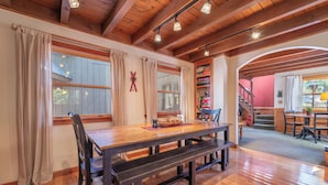 A dining area with a wooden table and benches, large windows with curtains, and a staircase leading to another room.
