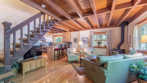 A spacious living area with a wooden staircase, a sofa, a TV, and a kitchen in the background.