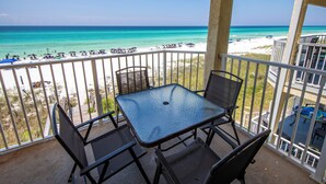 A balcony with a glass table and chairs overlooking a beach with umbrellas and people.