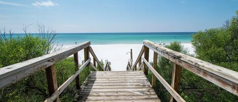 A wooden boardwalk leading to a beach with clear blue water and a sandy shore.
