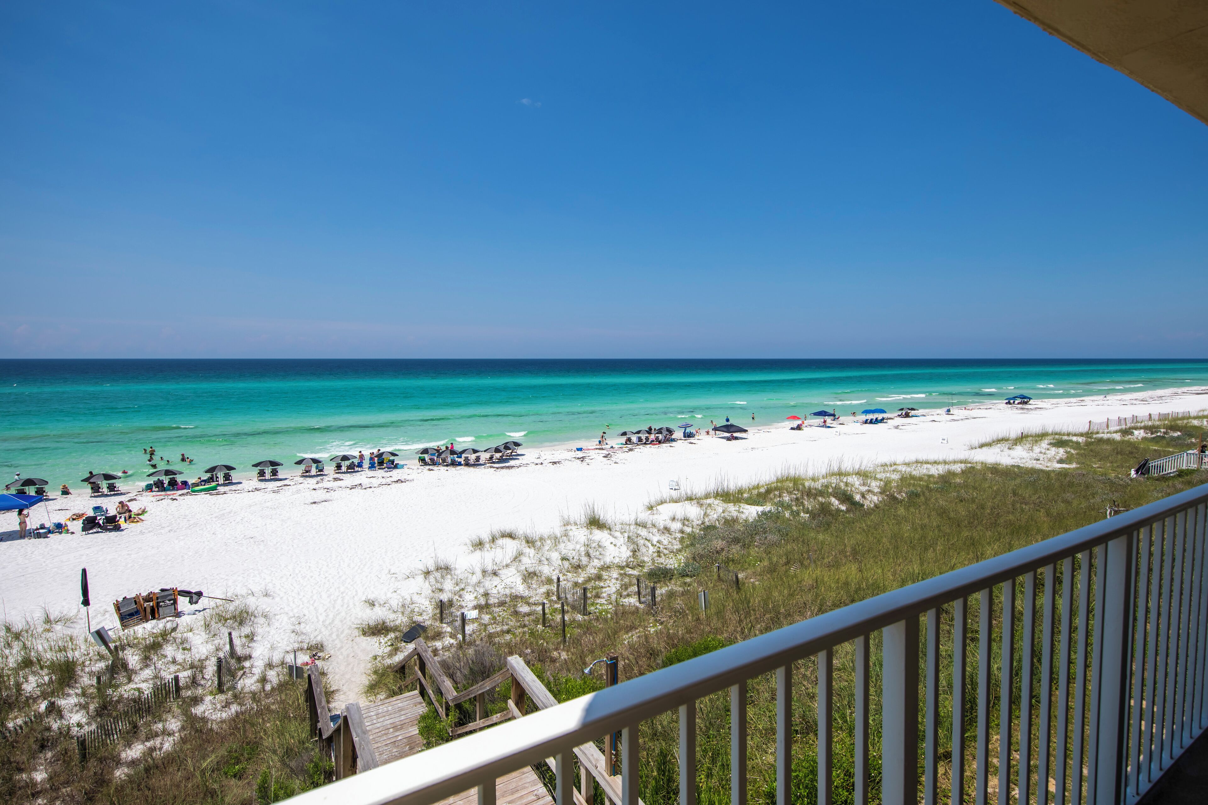 A beach with a wooden deck and a railing, overlooking a sandy beach with umbrellas and beach chairs.