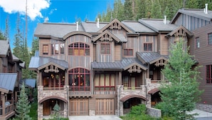A large, multi-story log cabin with a stone foundation, multiple chimneys, and a prominent front porch. - Base Camp 480 | Mountain Townhome | On The Slopes | Private Hot Tub (Winter Park)