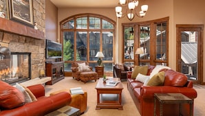A spacious living room with a stone fireplace, leather sofas, and large windows offering a view of a mountain lodge. - Base Camp 480 | Mountain Townhome | On The Slopes | Private Hot Tub (Winter Park)