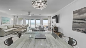 A modern dining area with a marble table, surrounded by chairs, in a room with a large window overlooking the ocean.
