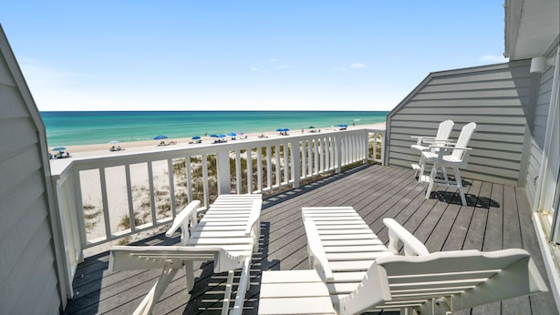 A wooden deck with white Adirondack chairs overlooking a beach with umbrellas and people.