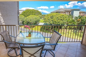 A balcony with a glass table and chairs, overlooking a residential area with houses and greenery.