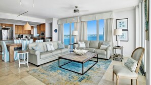A spacious living room with a large window offering a view of the sea, a kitchen with wooden cabinets, and a dining area with striped chairs.