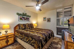A bedroom with a bed, bedside lamps, a ceiling fan, a window with blinds, and a wall-mounted TV.