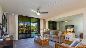 A spacious living room with a large sliding glass door leading to an outdoor patio, a ceiling fan, a sofa, a coffee table, and a wicker chair. - Kamaole Sands #10-111  Ocean View, Inner Court, Ground Floor, Close to Beach (Kihei)