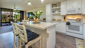 A modern kitchen with white cabinetry, a central island with seating, a microwave, oven, and a dining area with chairs and a table. - Kamaole Sands #10-111  Ocean View, Inner Court, Ground Floor, Close to Beach (Kihei)