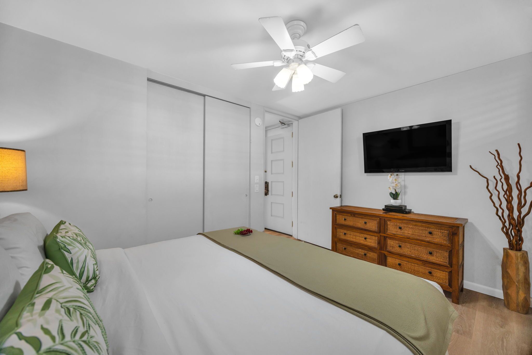 A bedroom with a bed, a wooden dresser, a ceiling fan, a TV, and a door.