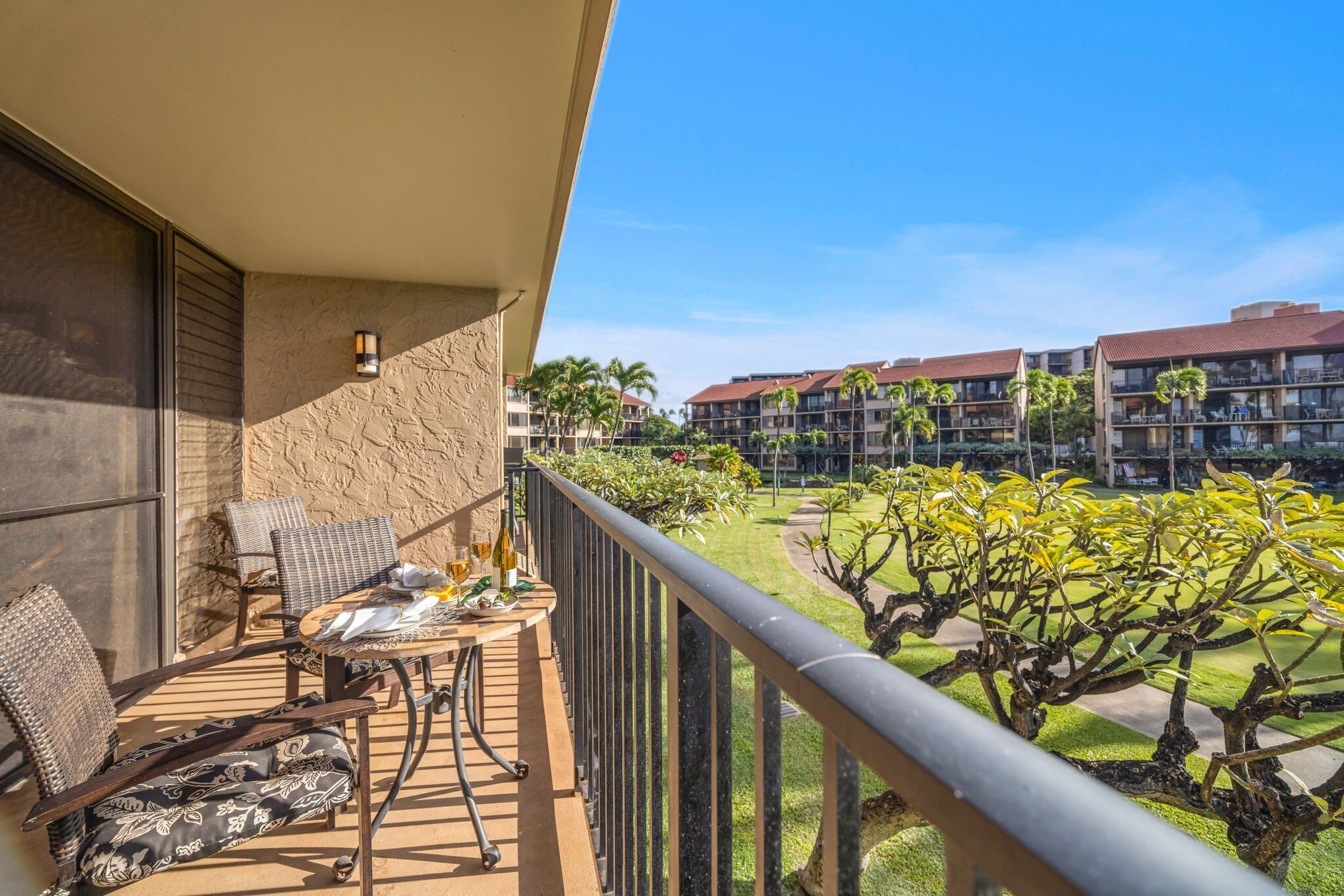 A balcony with wicker furniture, a table with a bottle and glasses, and a view of a landscaped garden with palm trees and a building complex in the background.