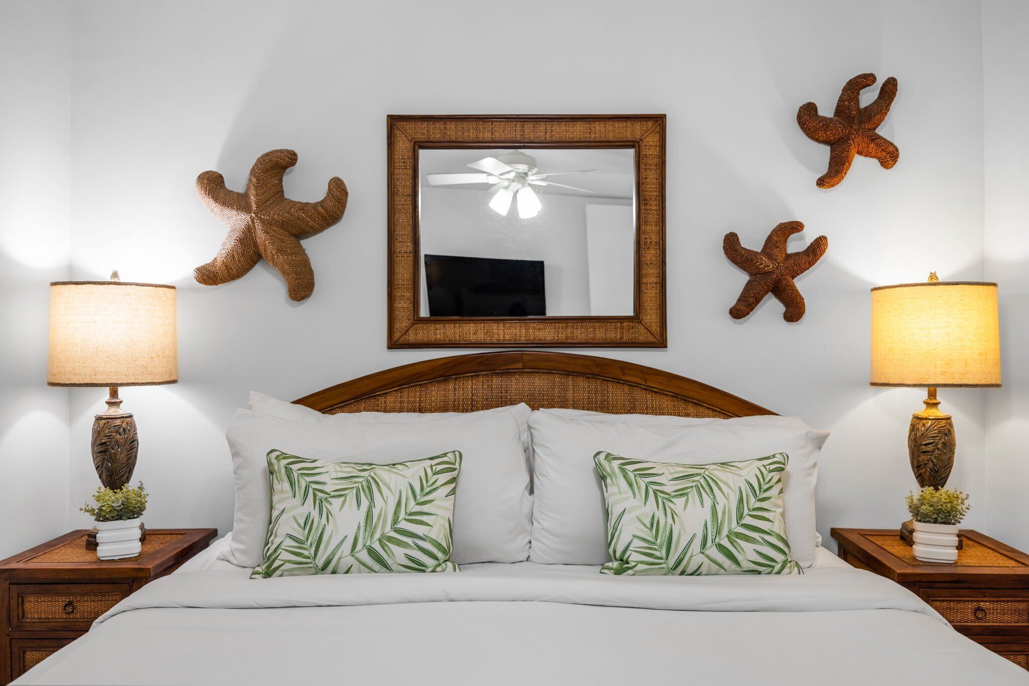 A neatly arranged bedroom with a bed, two nightstands with lamps, a ceiling fan, and wall decorations including starfish and a mirror.