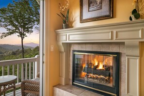 A fireplace with a fire, a wicker chair, and a balcony with a view of the mountains.
