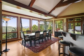 A dining area with a wooden table and chairs, large windows with a view of greenery, and a bar area with stools.