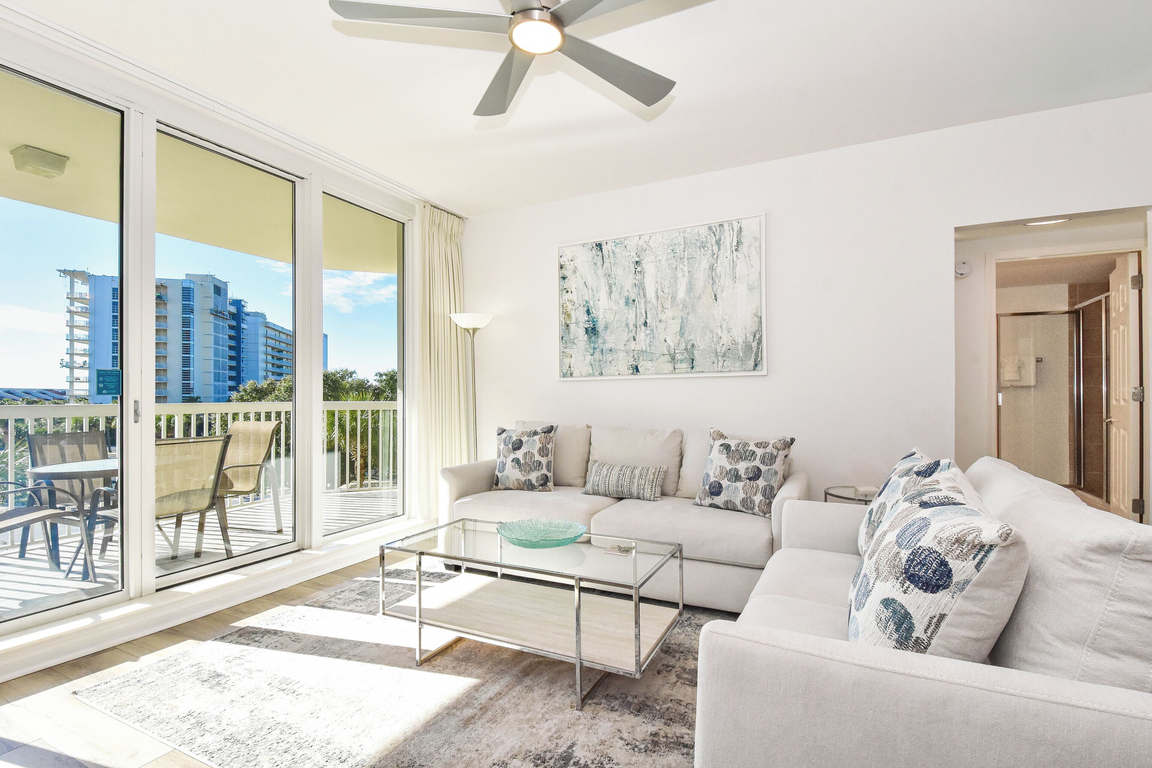 A modern living room with a glass coffee table, a sofa with patterned cushions, and a view of a balcony with outdoor furniture.