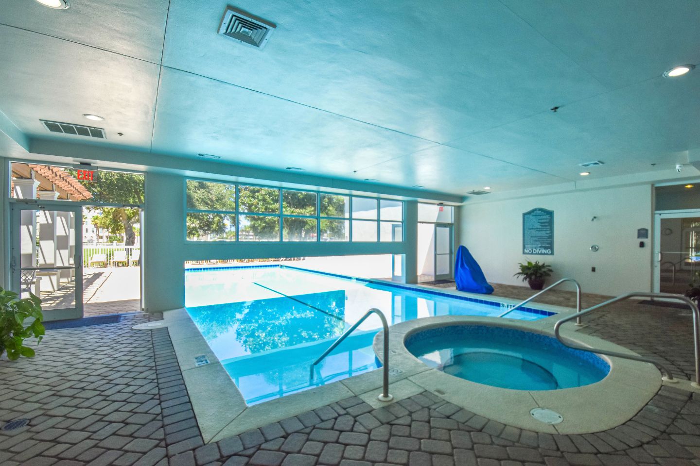 An indoor swimming pool with a clear view of the outside, a hot tub, and a poolside area with a covered seating area.