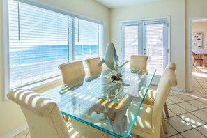 A dining area with a glass table and chairs, a view of the ocean through large windows, and a door leading outside.