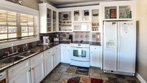 Une cuisine avec des meubles blancs, une cuisinière, un réfrigérateur et une fenêtre avec des stores.