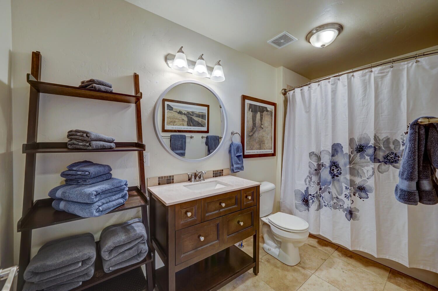 Une salle de bain avec un meuble-lavabo en bois, un miroir rond, des toilettes et un rideau de douche orné de motifs floraux.