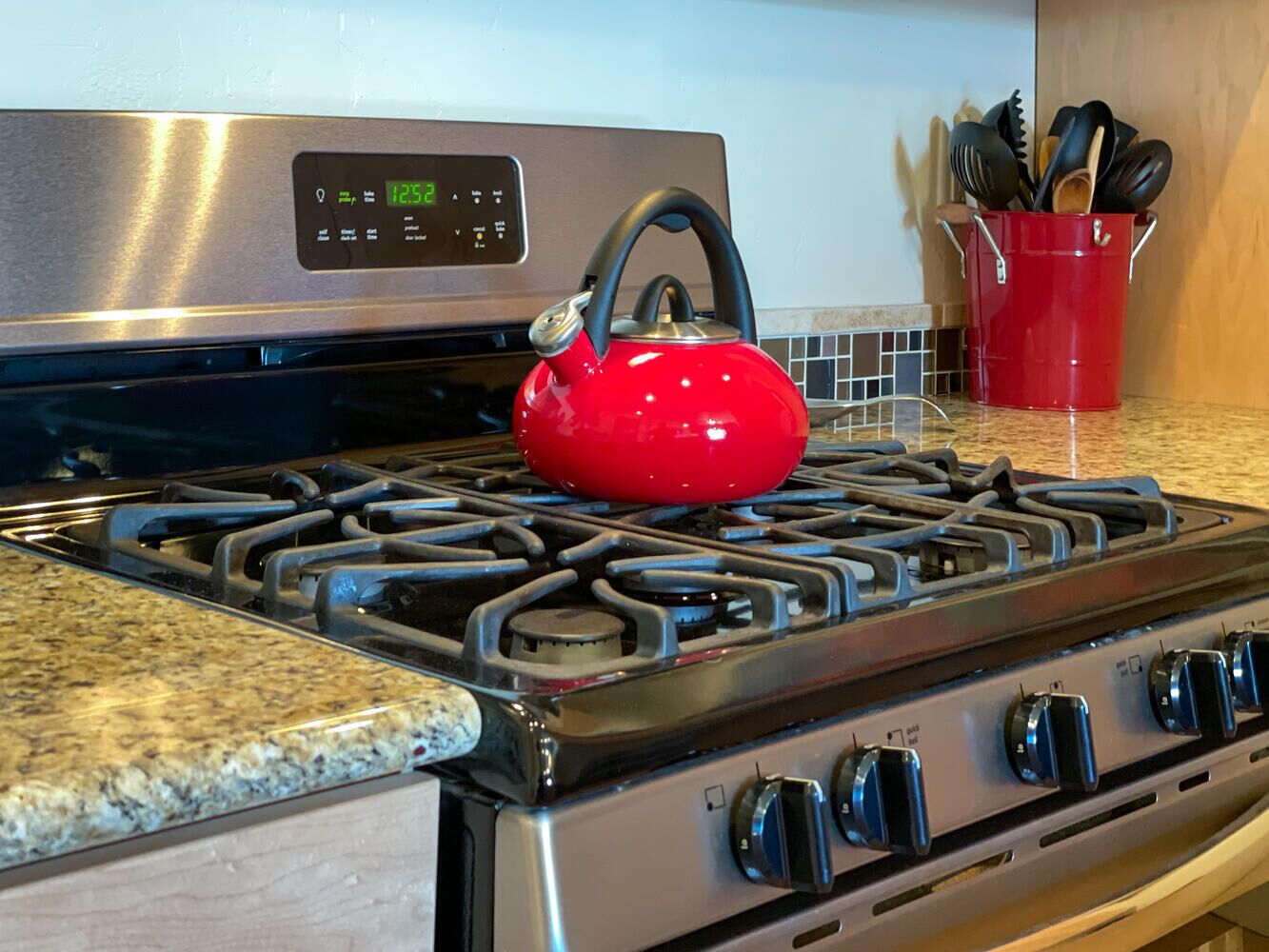 A kitchen with a stainless steel stove, a red kettle, and a red bucket with utensils.