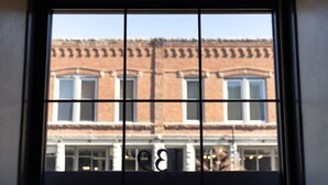 Una ventana con un diseño de cuadrícula, a través de la cual se ve un edificio con múltiples ventanas y fachada de ladrillo.