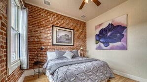 Un dormitorio con pared de ladrillo, una cama grande con ropa de cama gris, un cuadro en la pared y una obra de arte floral de gran tamaño.