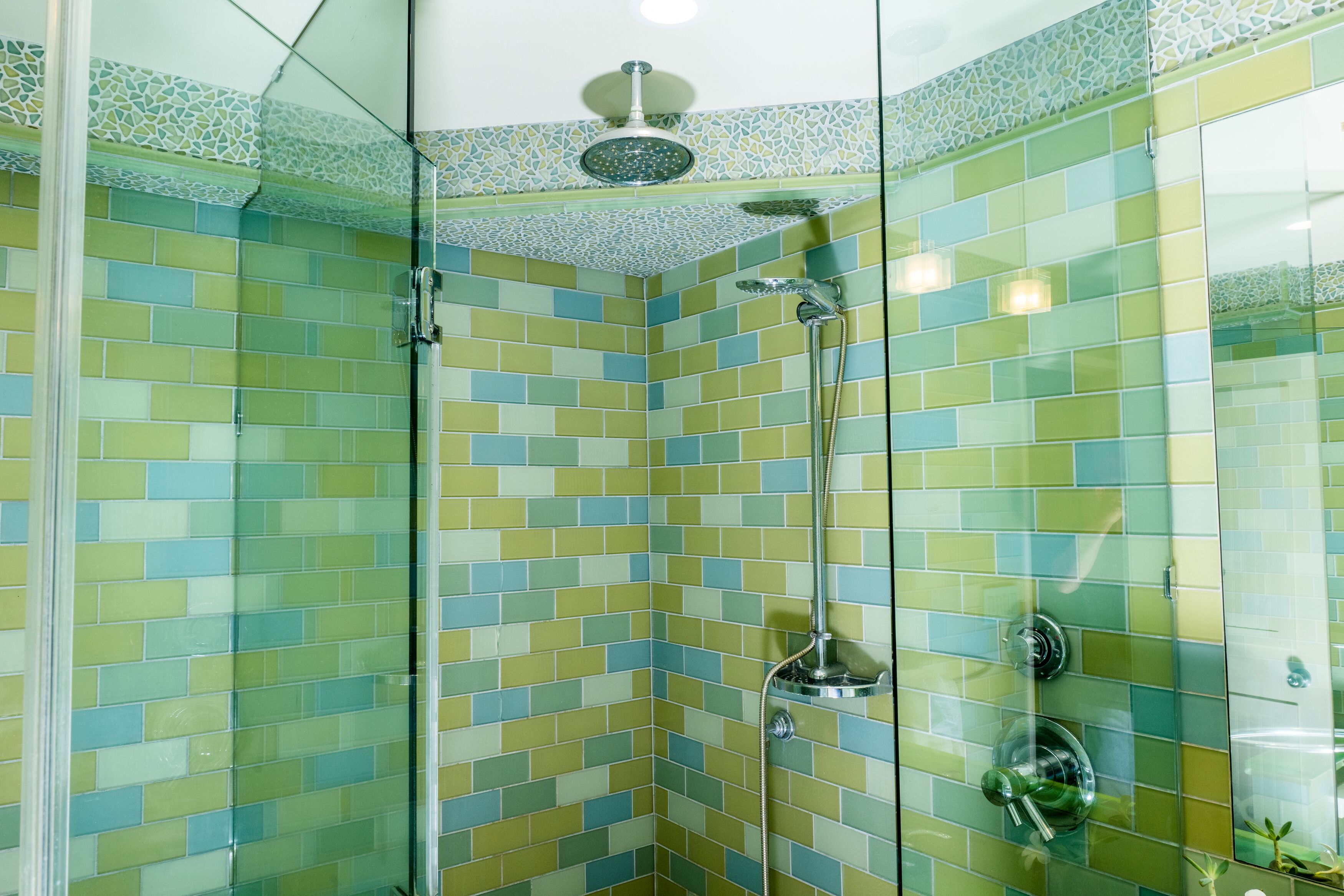Une douche d’angle avec une porte en verre, des carreaux de mosaïque verts et jaunes, et un pommeau de douche à main.