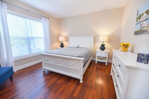 Een slaapkamer met een houten vloer, een wit bed met hoofdeinde, twee nachtkastjes met lampen, een dressoir en een raam met gordijnen.