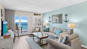 A living room with a sofa, armchairs, a coffee table, and a TV. There are two lamps, a fish-shaped wall decoration, and an anchor decoration. A window with a view of the ocean is visible. - 🌴Views~Pool~Tennis~2BR Hol Surf & Racquet Club 710 (Destin)