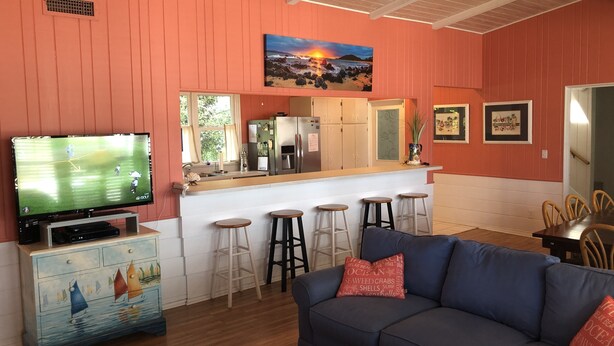 Een woonkamer met een televisie, een bank, een salontafel en een keukenhoek met een bar en barkrukken.