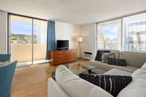 Una sala moderna con sofá, una mesa de centro, un televisor sobre un mueble de madera y vistas de la ciudad a través de grandes ventanales.