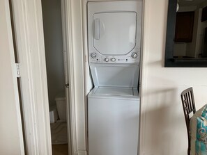 A compact, white washing machine in a narrow hallway with a door leading to a bathroom.