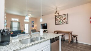 A modern kitchen with a granite countertop, stainless steel sink, and a dishwasher. There is a dining area with a wooden table and bar stools. The room has a ceiling fan, pendant lights, and a wall-mounted television. - Pet-Friendly Beach Escape | Pool Access & Walk to Beach (Corpus Christi)