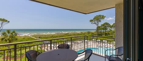 A balcony with a table and chairs, overlooking a beach and a swimming pool.