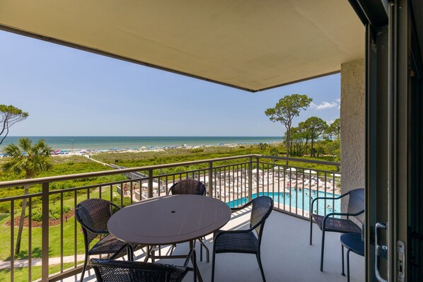 A balcony with a table and chairs, overlooking a beach and a swimming pool.