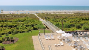 Un hotel frente a la playa con un patio que cuenta con mesas y sillas al aire libre y sombrillas, un camino que conduce a la playa y vistas al océano.