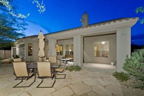 Una casa moderna con patio, che include un tavolo e sedie in pietra, un ombrellone e vista sull'interno tramite ampie finestre.
